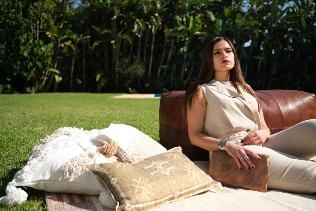 Femme adossée à un pouf vendu par Mazen portant les bijoux et le sac Mazen ainsi que des coussins posés au sol sur un tapis